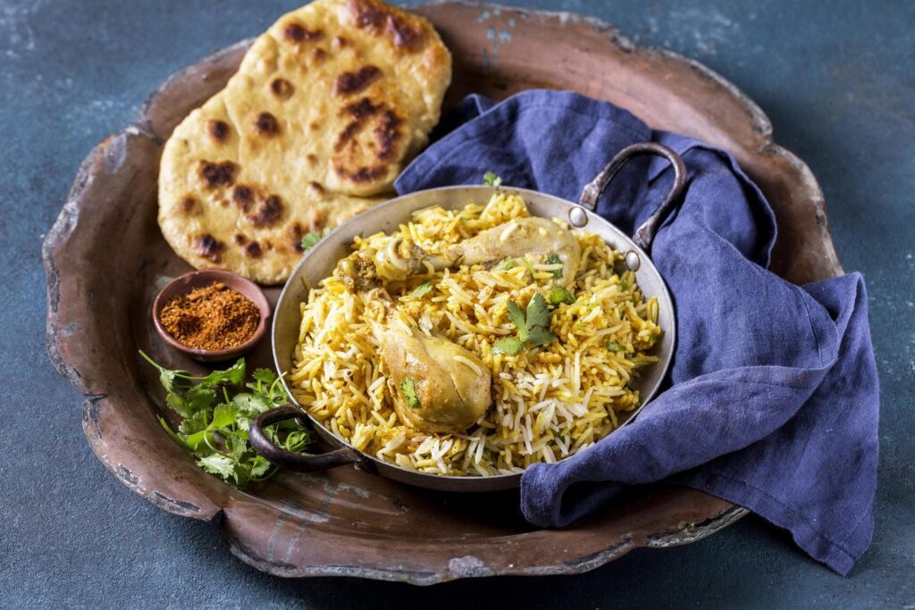 Top-angle view of golden chicken biryani cooked with EatMe parboiled rice, garnished with fresh cilantro, served with naan bread and spices on a rustic tray. 俯角拍攝以益米預熟米烹調的金黃雞肉印度香飯，飾以新鮮香菜，搭配烤餅與香料盛於復古托盤中。