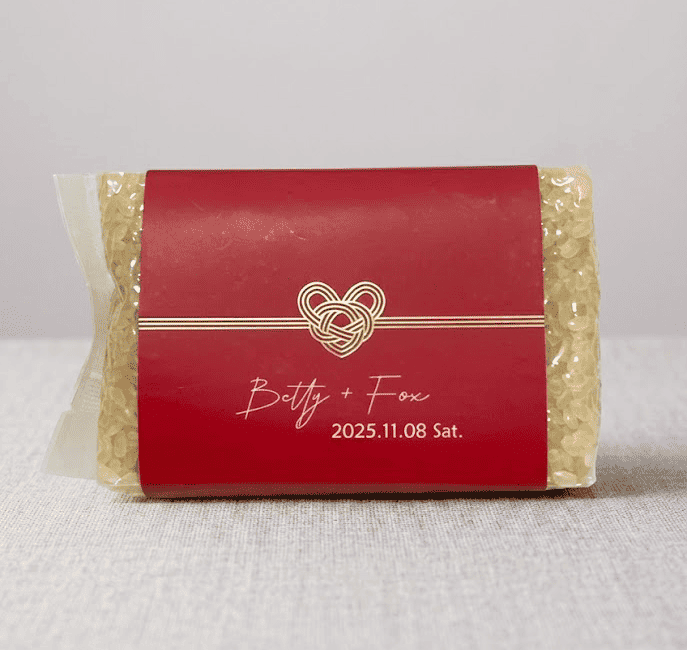 A transparent package of EatMe parboiled rice sealed with a red custom wedding label printed with a gold knot heart design and the couple's names "Betty + Fox, 2025.11.08 Sat.", presented as a meaningful wedding favor gift. 透明包裝的益米預熟米，貼有紅色客製婚禮標籤，印有金色結心圖案及新人姓名「Betty + Fox, 2025.11.08 Sat.」，作為富有意義的婚禮伴手禮。