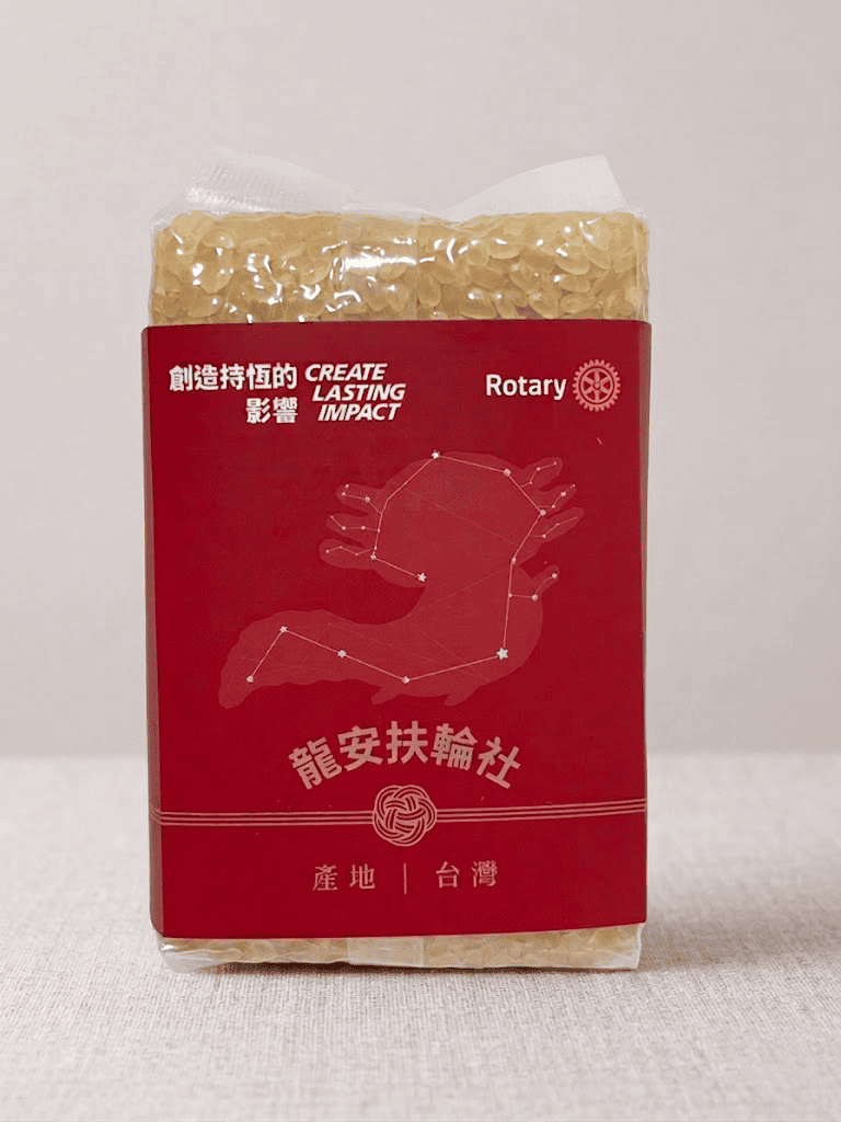 Close-up of a transparent EatMe parboiled rice package wrapped with a red custom label featuring the Rotary International logo, "Create Lasting Impact" bilingual slogan, Long-An Rotary Club name, and "Product of Taiwan" designation. 透明包裝益米預熟米特寫，外貼紅色客製標籤，印有國際扶輪 Logo、「創造持恆的影響」中英雙語口號、龍安扶輪社名稱及「產地：台灣」標示。
