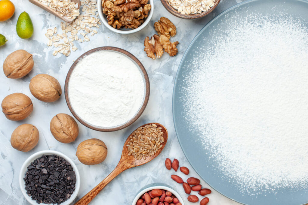 Top-view flatlay of white flour in a bowl surrounded by walnuts, rolled oats, sunflower seeds, peanuts, and chocolate chips on a marble surface — representing a variety of whole grain and nut ingredients compared to refined carbs, in the context of choosing healthier staple foods. 俯拍大理石桌面擺放白麵粉碗,周圍散布核桃、燕麥片、葵花籽、花生與巧克力碎片,呈現全穀物與堅果食材與精製碳水化合物的對比,說明選擇更健康主食的概念。