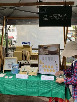 Eatme parboiled rice market stall (booth C03) at a Taiwan outdoor fair, featuring packaged rice products priced at 180 NTD, promotional posters highlighting 8 key benefits, and a QR code for online ordering. 益米預熟米台灣戶外市集攤位（C03號），展示定價180元台幣的包裝預熟米、強調8大優點的宣傳海報，以及線上訂購QR碼。
