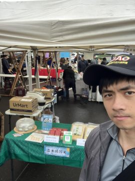 Eatme brand representative standing in front of an outdoor market booth displaying EatMe parboiled rice products, packaged goods, and pricing signs at a Taiwan local fair. 益米預熟米品牌人員站於台灣戶外市集攤位前，展示益米預熟米產品、包裝商品與價格標示。